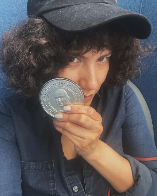 Adult female with curly hair and a black baseball cap holds a coin with a portrait in an indoor setting, wearing a blue denim shirt.