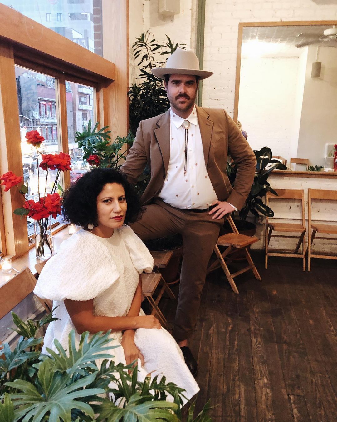 Man and woman in formal wear pose indoors surrounded by plants, a window, and wooden chairs in a candid, eye-level portrait.