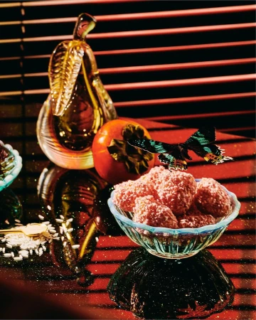 Still life photo of crystal glass bowl of pink coconut candies, glass pear, persimmon, and black butterfly on reflective surface.