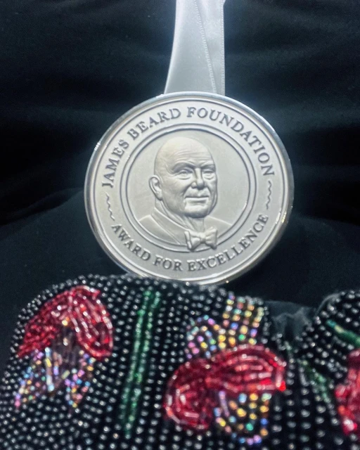 James Beard Foundation Award for Excellence medallion displayed on black fabric background with an embroidered floral bag at the bottom.