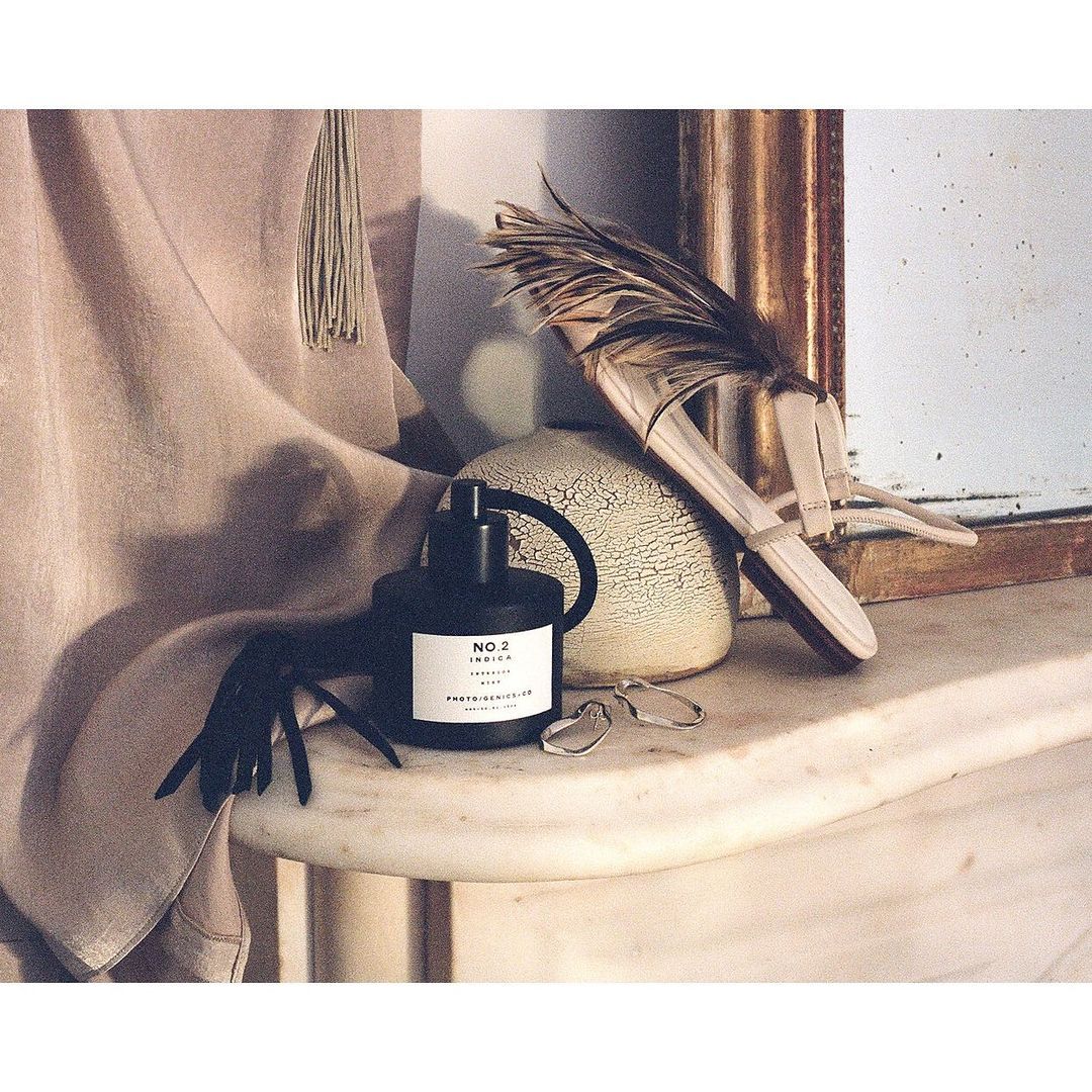 Still life photo of a black perfume bottle, feather duster, and cream-colored vase on a white marble table with a gold-framed mirror.
