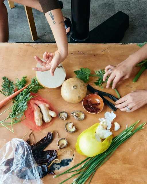 Overhead shot of vegetables, mushrooms, chives, peppers, and garlic bulbs arranged on a wooden table, with two female hands reaching in.