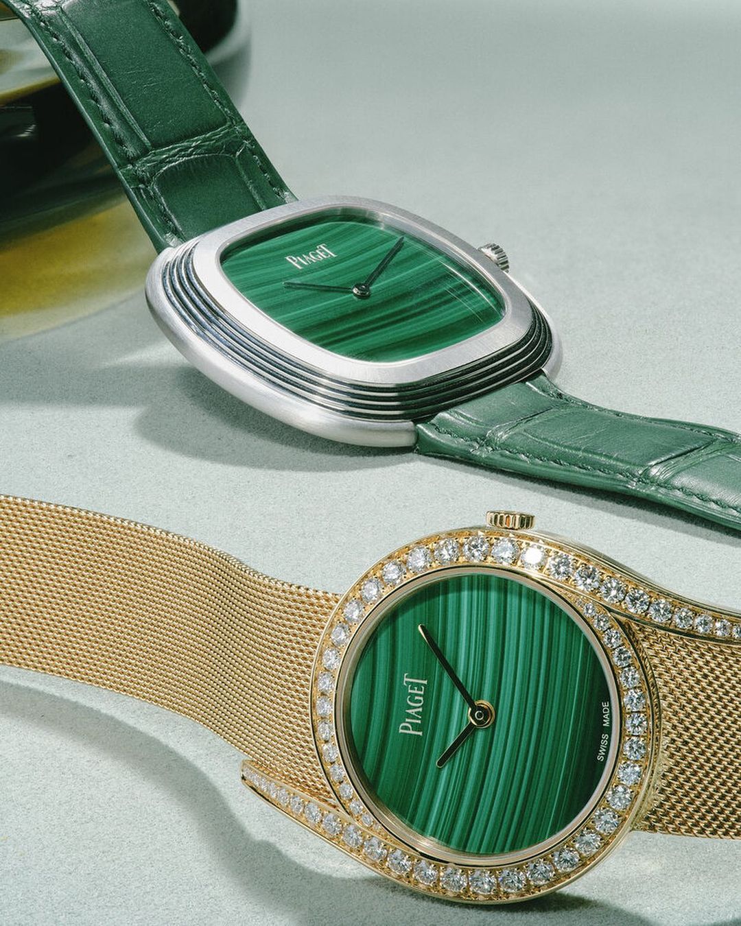 Two Piaget watches, one silver with a green leather strap and one gold with a diamond bezel, are displayed against a soft grey background.