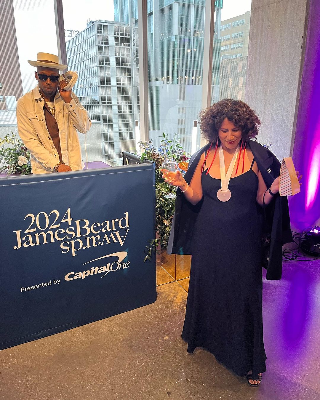 Two people at the 2024 James Beard Awards in an indoor setting, with city buildings visible through a window in the background.