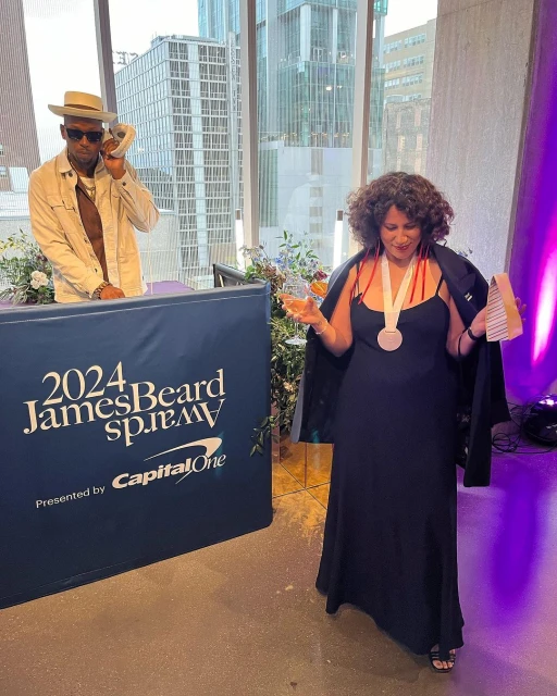 Two people at the 2024 James Beard Awards in an indoor setting, with city buildings visible through a window in the background.