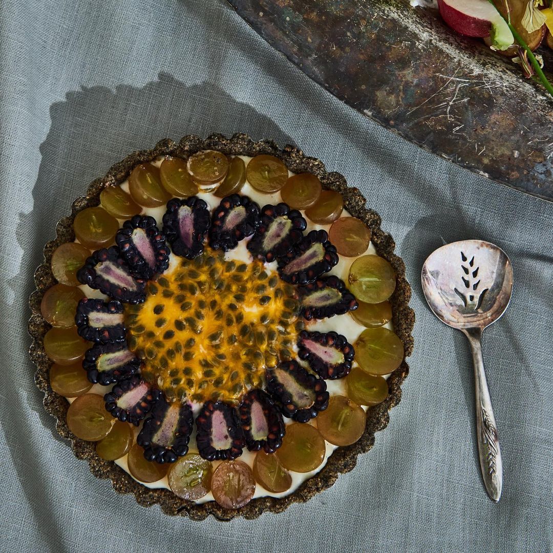 Overhead shot of a fruit tart with blackberries, gooseberries, and passionfruit, set on a grey linen background next to a silver spoon.