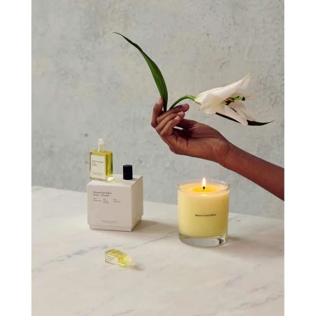 Perfume bottle, candle, and white flower on marble table with a woman's hand holding the flower against a light wall backdrop.