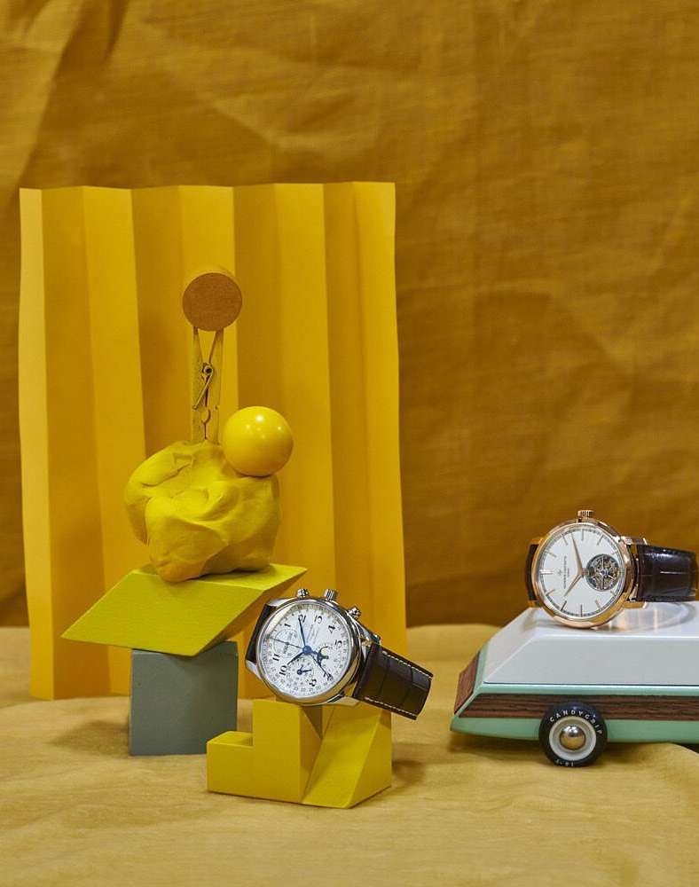 Two wristwatches displayed on a miniature trailer and geometric forms against a yellow backdrop in a studio setting with soft lighting.