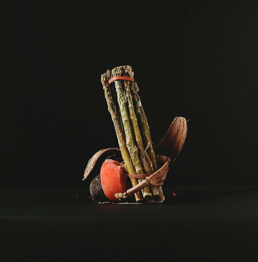 Close-up still life of green asparagus spears bundled with an orange band, featuring an avocado half and dried botanical elements against a black background.