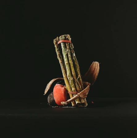 Close-up still life of green asparagus spears bundled with an orange band, featuring an avocado half and dried botanical elements against a black background.