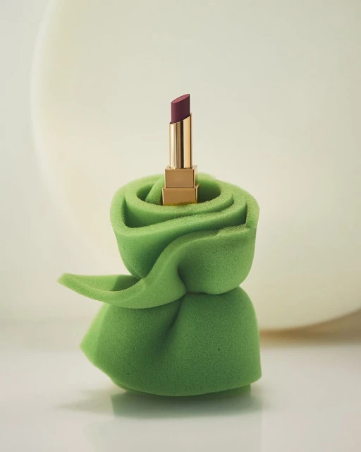 Still life of a single lipstick standing atop a green sponge, set against a backdrop of soft, muted colors. Studio shot.