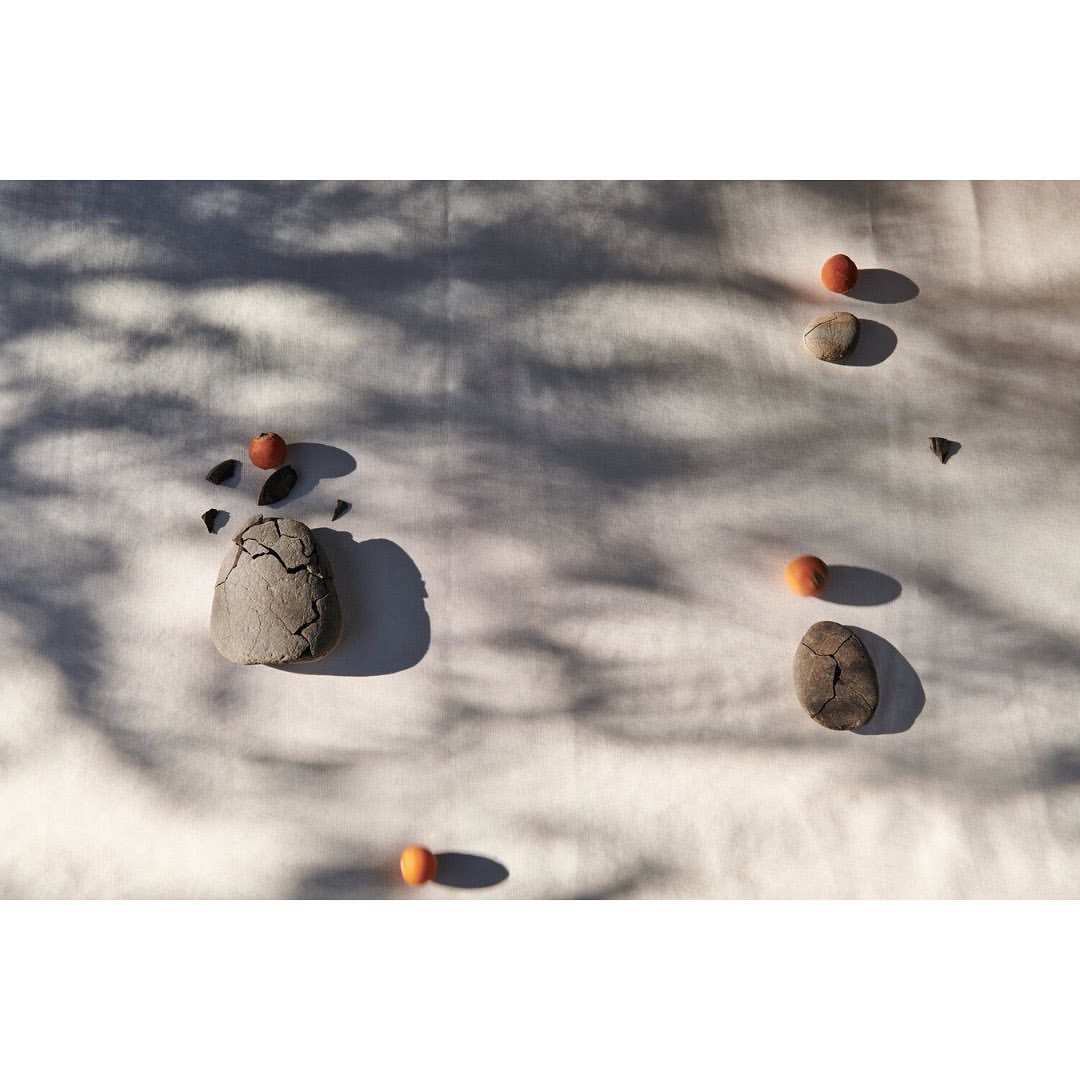 Still life featuring stones and persimmons scattered on a white cloth, with dappled shadows creating a minimalist and serene composition.