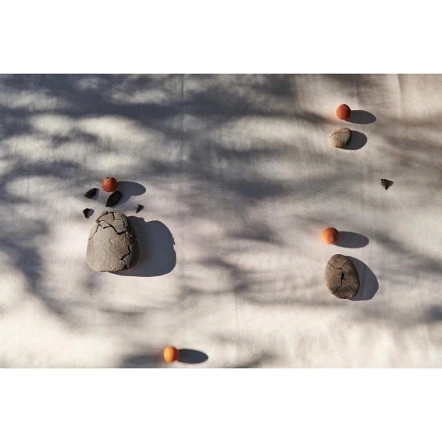Still life featuring stones and persimmons scattered on a white cloth, with dappled shadows creating a minimalist and serene composition.
