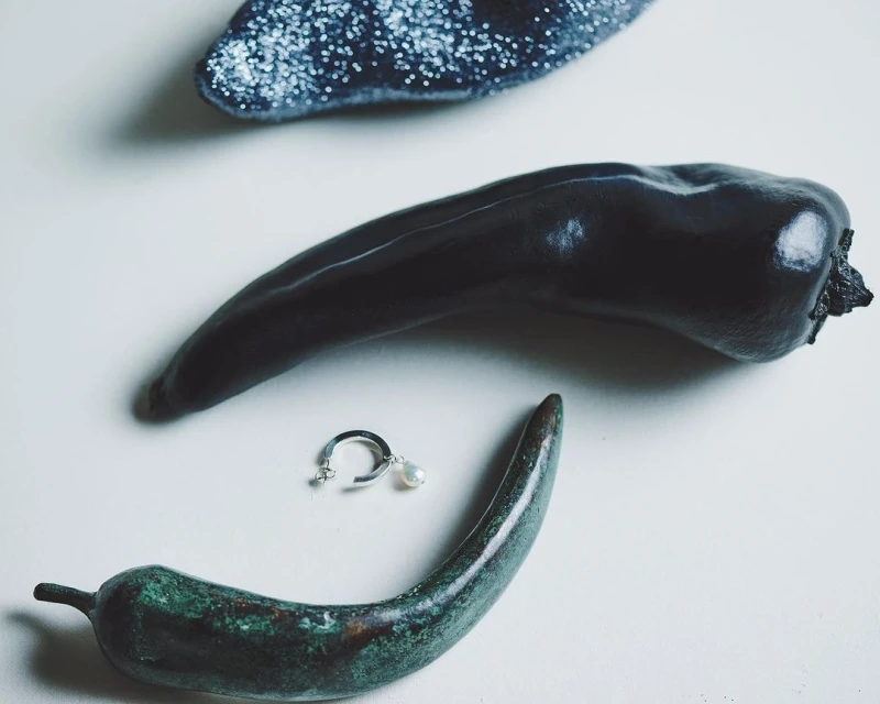 Still life of black and green peppers and a silver earring with pearl drop on a white surface in a studio setting.