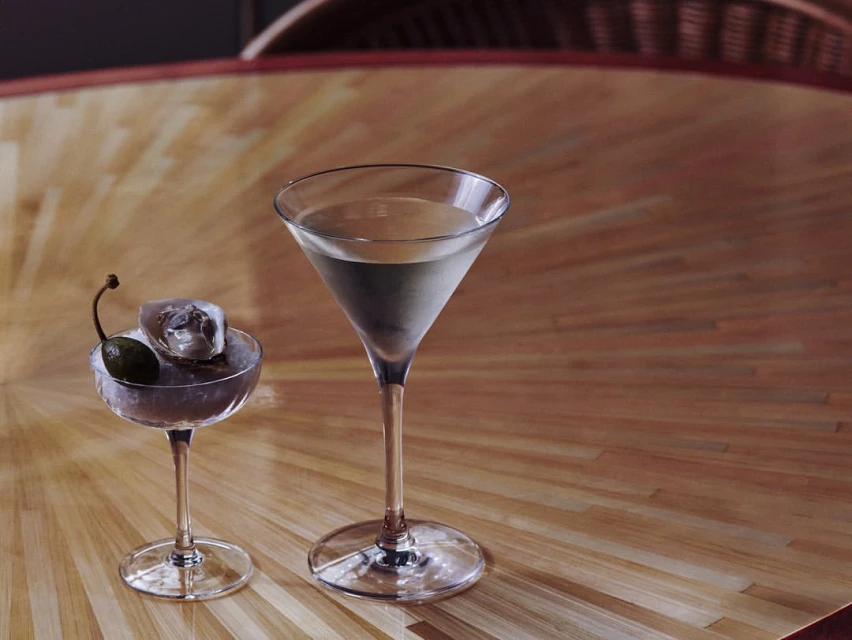 Two martini glasses, one with a cocktail olive and an ice cube, sit on a wood table with a radial pattern in a dimly lit interior.