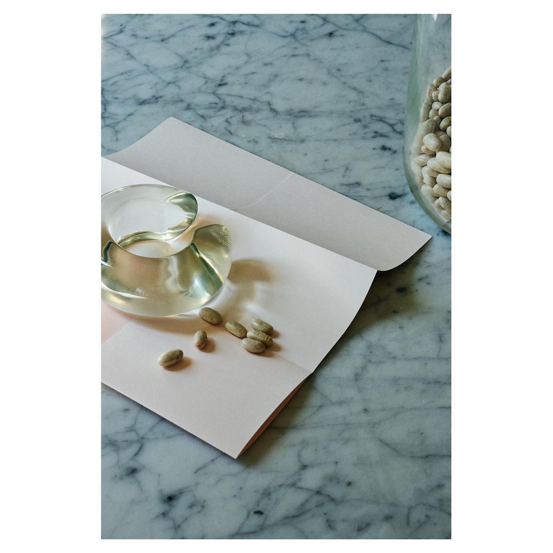 Still life photo of a clear glass cup and saucer, pistachios, and a glass jar of white beans on a marble surface with soft, natural light.