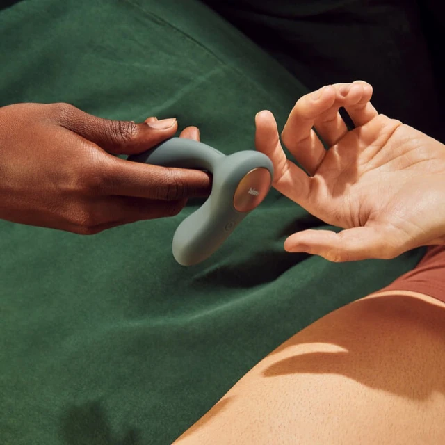 Two hands, one with dark skin and one with fair skin, hold a gray curved vibrator against a dark green fabric background.