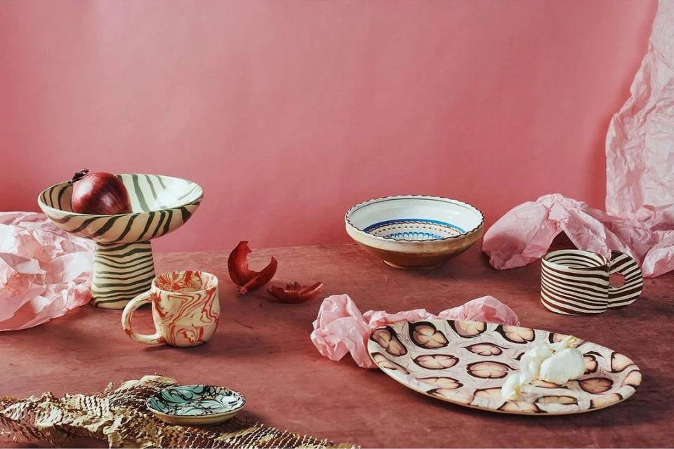Still life photograph of arranged ceramic dishes including bowls, mugs, and plates in a pink-toned studio setting with natural lighting.