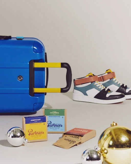 Still life shot of blue luggage, pair of high-top sneakers, ornament balls, and boxes of Partner's Coffee on a white backdrop.