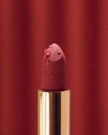 Close-up of a pink Gucci lipstick bullet with water droplets against a dark red backdrop