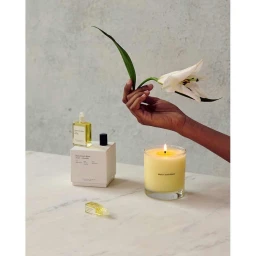 Hand holding a white lily flower above a lit candle and perfume bottles on marble