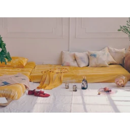 Low floor bed covered in yellow bedding with patterned throw pillows and red shoes on a white floor in a bright room.