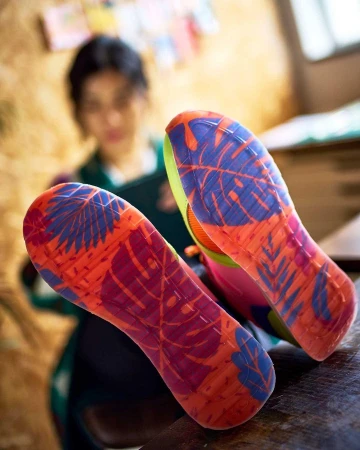 Close-up on the colorful soles of a pair of sneakers with a woman seated in the background, partially out of focus, creating a lifestyle aesthetic.
