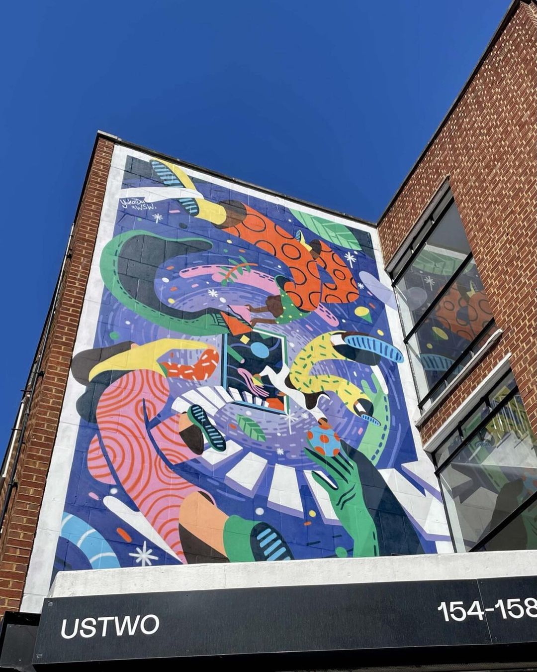 Eye-level shot of a vibrant mural with abstract shapes, figures, and colors on the exterior brick wall of an urban building under blue sky.