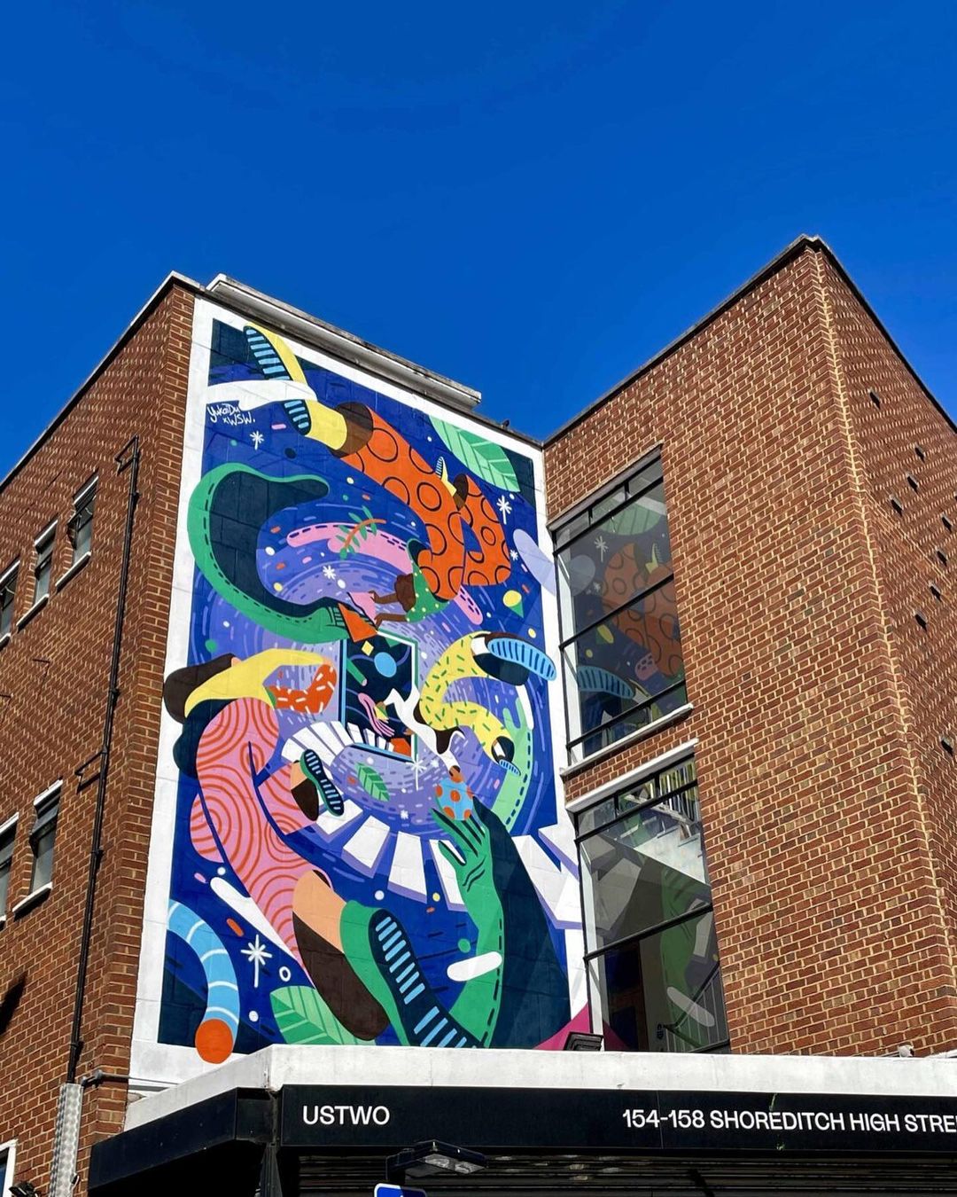 Exterior shot of a red brick building featuring a large, colorful mural with abstract figures and shapes under a clear blue sky.