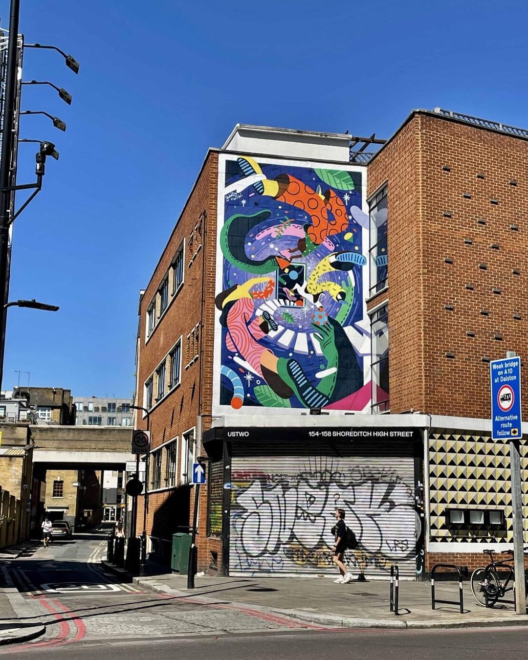 Outdoor street art mural on brick building with colorful abstract patterns, pedestrian, sidewalk, graffiti-covered storefront, sunny day.