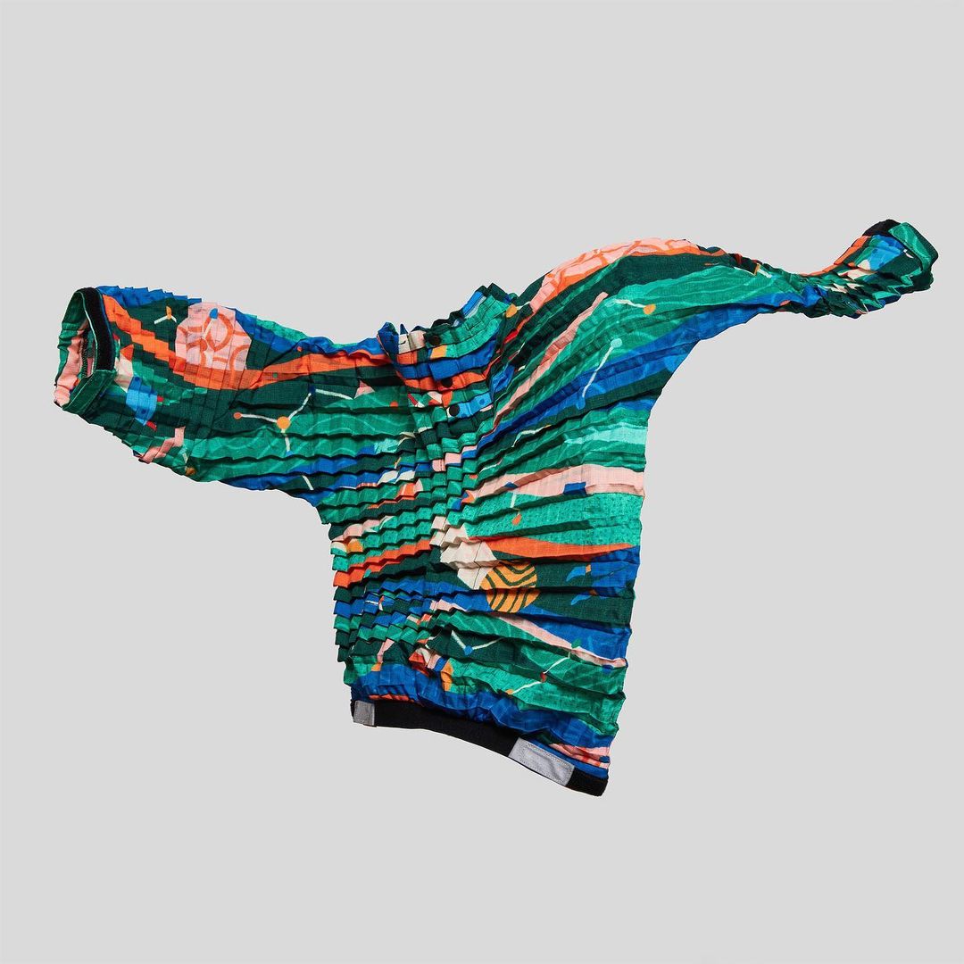 Wrinkled, pleated short-sleeved top with green, blue, orange, and pink graphic pattern, and a black collar and hem, floating in studio
