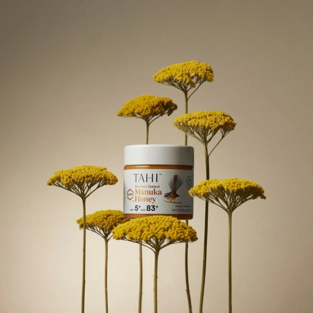 Still life shot of a jar of Tahi Manuka Honey surrounded by yellow yarrow flowers against a light beige background in a studio setting.