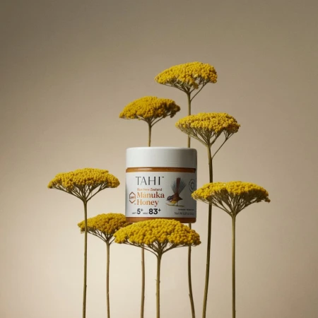 Still life shot of a jar of Tahi Manuka Honey surrounded by yellow yarrow flowers against a light beige background in a studio setting.