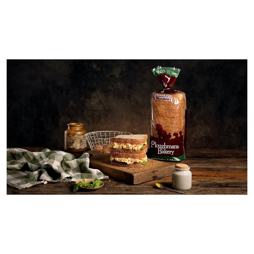 Three-layer sandwich on wooden board with bread bag, jar, and green cloth against dark background, in a studio food photography setting.