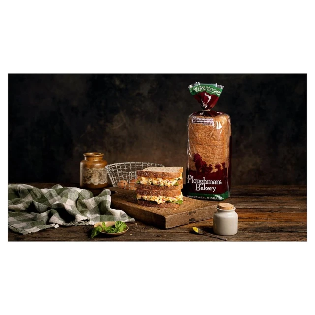 Three-layer sandwich on wooden board with bread bag, jar, and green cloth against dark background, in a studio food photography setting.