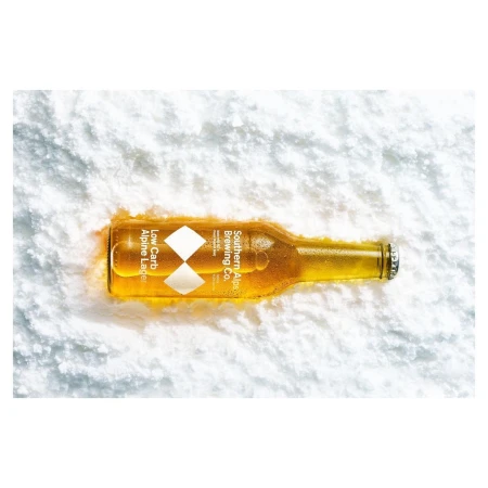 Close-up of a glass beer bottle lying horizontally on a bed of white granular snow. The bottle is amber-colored with branding.