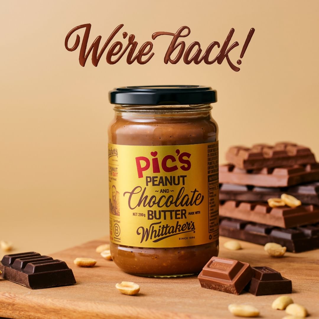 A jar of Pic's Peanut and Chocolate Butter sits on a wooden board with chocolate bars and peanuts against a tan backdrop.
