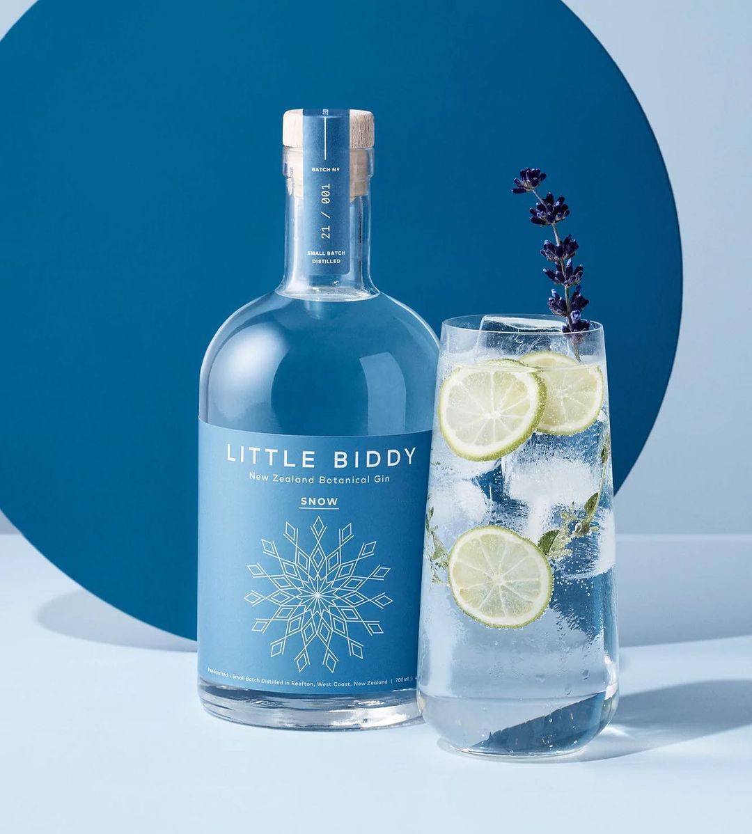 Studio shot of a gin bottle with lavender, beside a tonic water glass with lime garnish and ice, against a light blue backdrop.