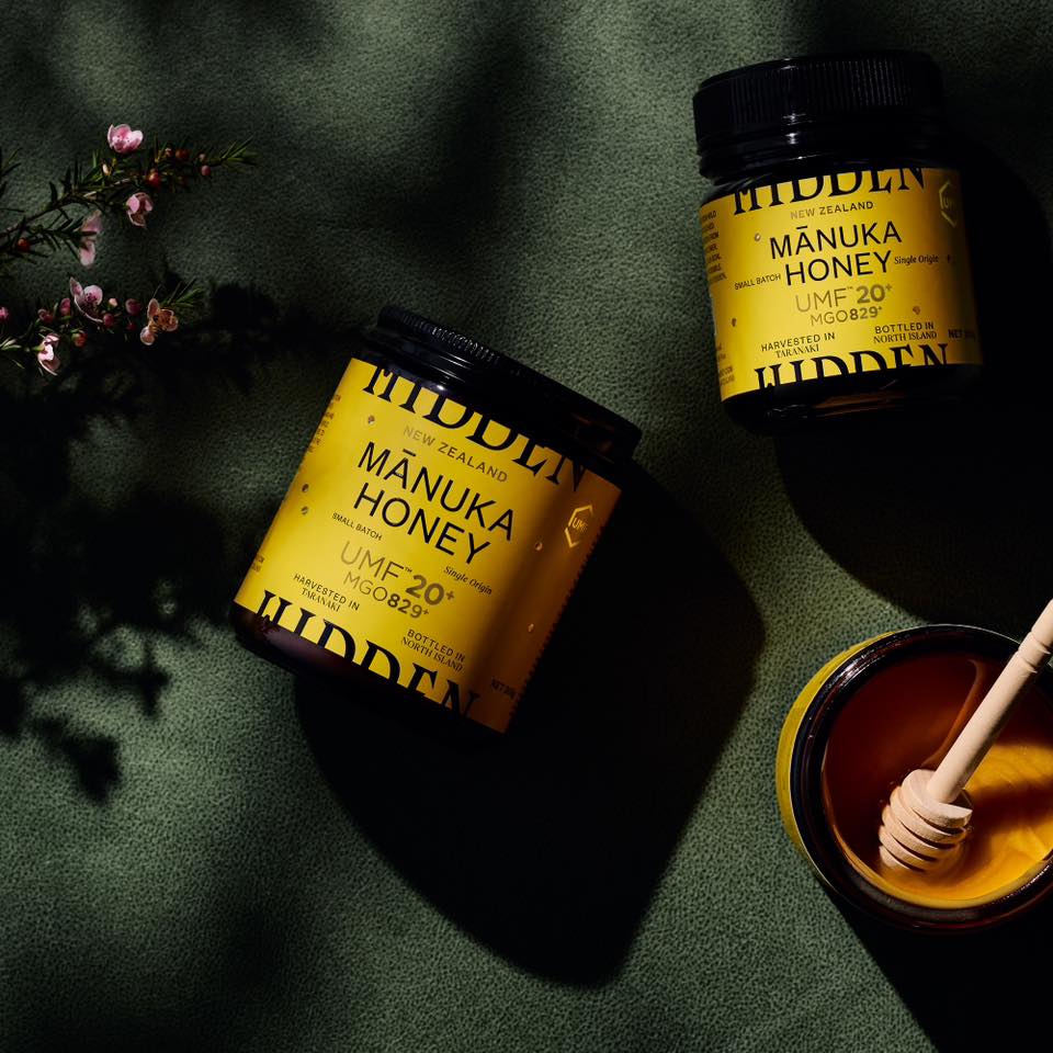 Overhead shot of two jars of New Zealand Manuka honey with a honey dipper and pink flowers against a textured green surface.