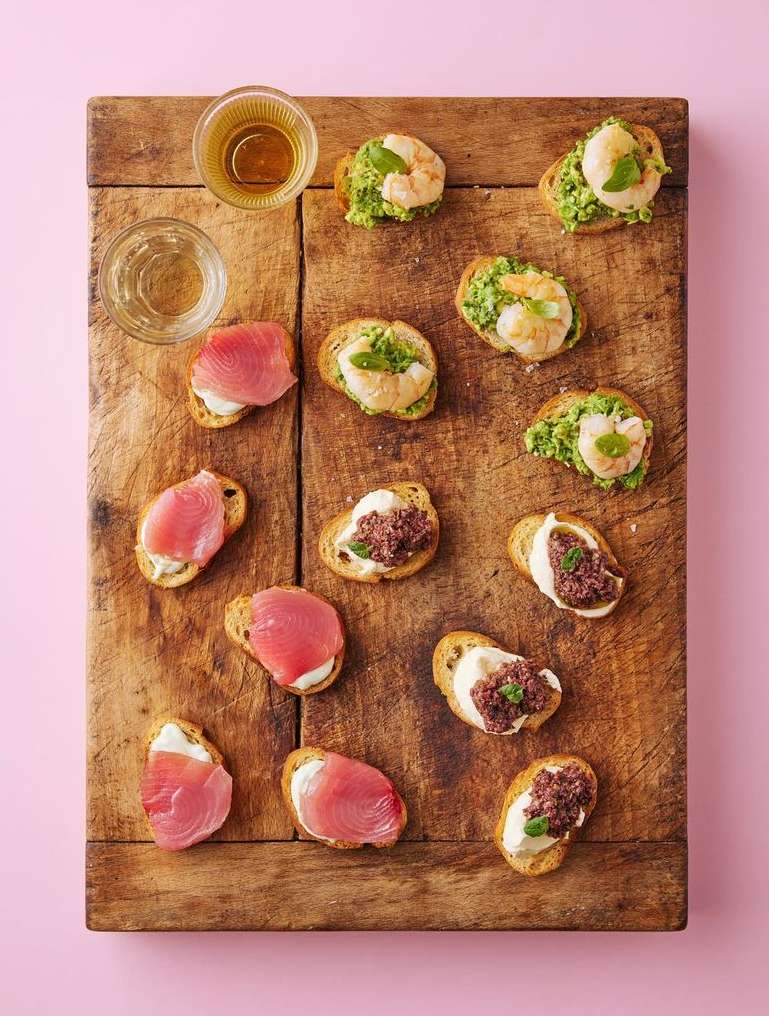 Overhead shot of tuna, shrimp, and olive bruschetta appetizers served with clear beverages on a wooden board against a pink background.