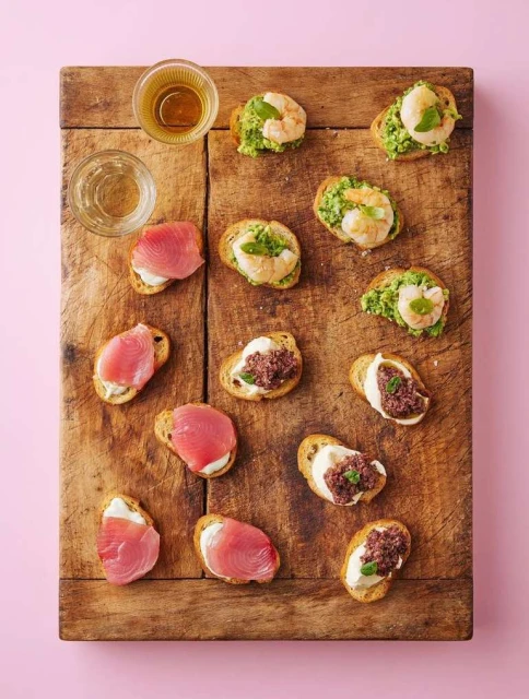 Overhead shot of tuna, shrimp, and olive bruschetta appetizers served with clear beverages on a wooden board against a pink background.