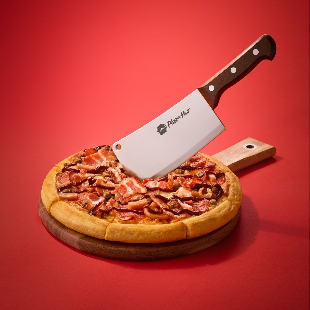 Overhead shot of a pizza topped with bacon and sliced meat, served on a wooden board, cut in half with a Pizza Hut cleaver.