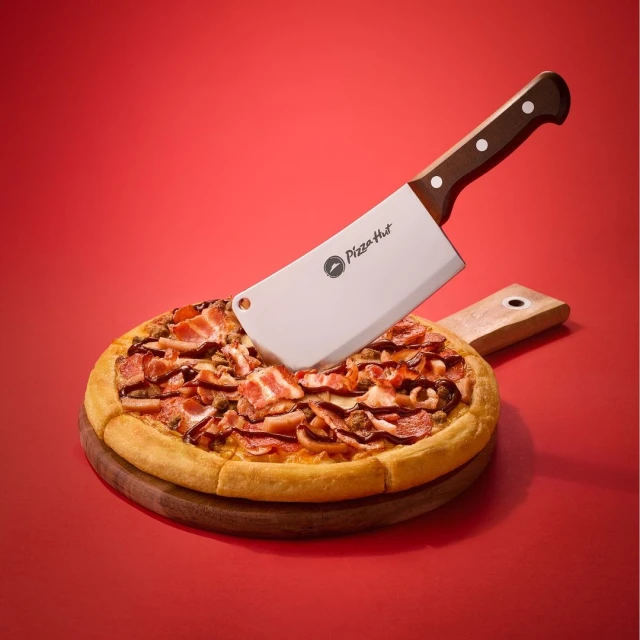 Overhead shot of a pizza topped with bacon and sliced meat, served on a wooden board, cut in half with a Pizza Hut cleaver.