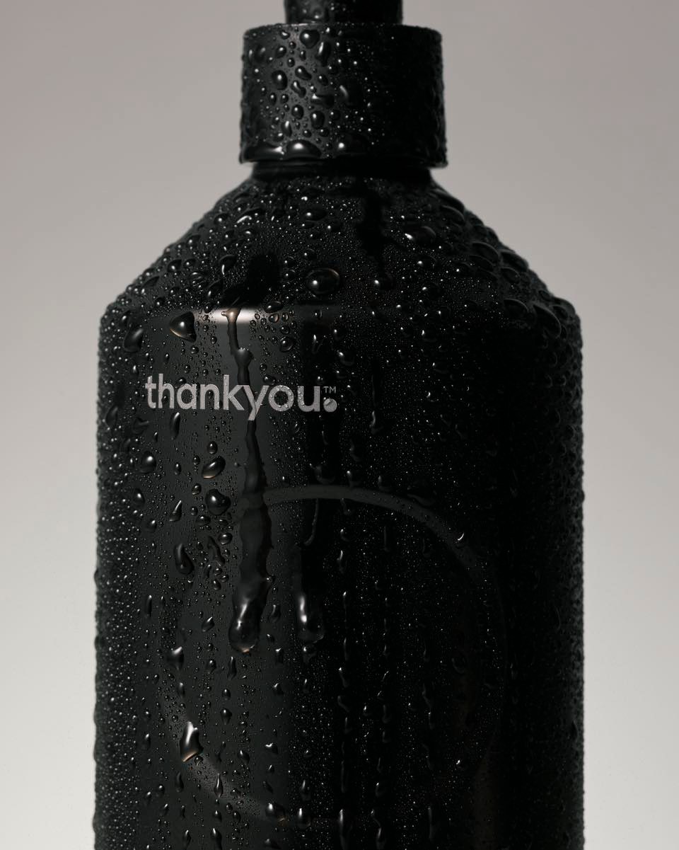 Close-up of a black plastic bottle covered in water droplets against a plain grey background, featuring the brand name 'thankyou.'.