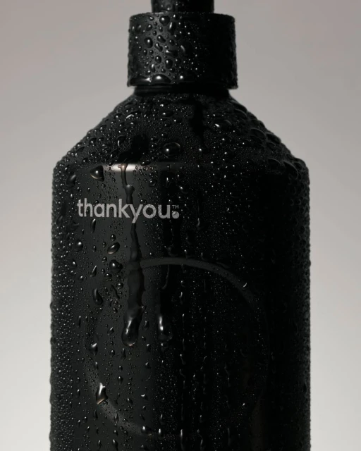 Close-up of a black plastic bottle covered in water droplets against a plain grey background, featuring the brand name 'thankyou.'.