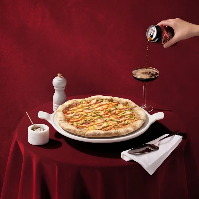 Overhead shot of pizza and soda served on a red tablecloth, with a salt shaker, glass, and can, indoors with studio lighting.