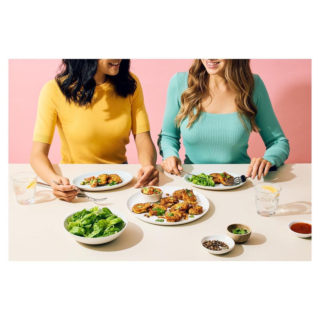 Two women eat healthy lunches together at a bright table with leafy greens, grains, and potatoes against a light pink background.