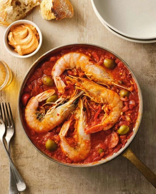 Overhead shot of shrimp cooked in tomato sauce in a copper pan, served with sliced bread, cream dip and cutlery on a beige table.
