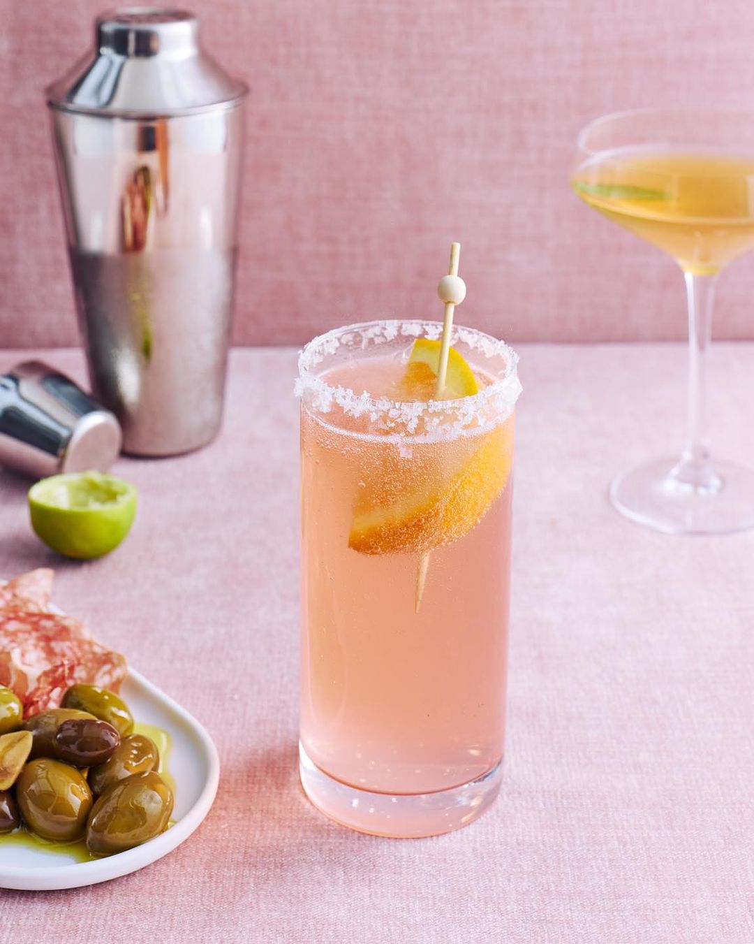 Tall cocktail glass with a salted rim and fruit garnish stands next to a shaker, lime half, and olives against a pale pink backdrop.