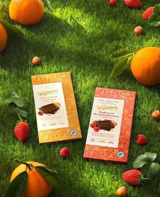Two Whittaker's chocolate bars surrounded by oranges, strawberries, and hazelnuts on a vibrant green grassy surface in bright light.